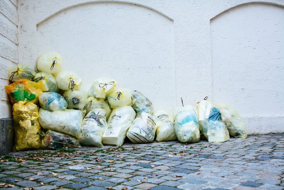 A collection of tightly packed rubbish bags, primarily yellow and transparent, rest on a cobblestone pavement against a plain white wall. Some of the bags are filled with plastic, paper, or other debris, with black ties sealing their openings. The bags are arranged in a somewhat haphazard manner, with a few leaning against each other and some overlapping. The environment appears to be an outdoor urban setting, possibly a back alley or side street, with natural daylight illuminating the scene. The textured cobblestones beneath the bags are uneven, with visible small stones and scattered leaves at their edges. This scene reflects typical private waste disposal arrangements where individuals or companies like Rubbish Clearance Marylebone handle bulky waste collection or rubbish removal, illustrating an alternative to traditional municipal waste services for disposing of household or commercial rubbish.