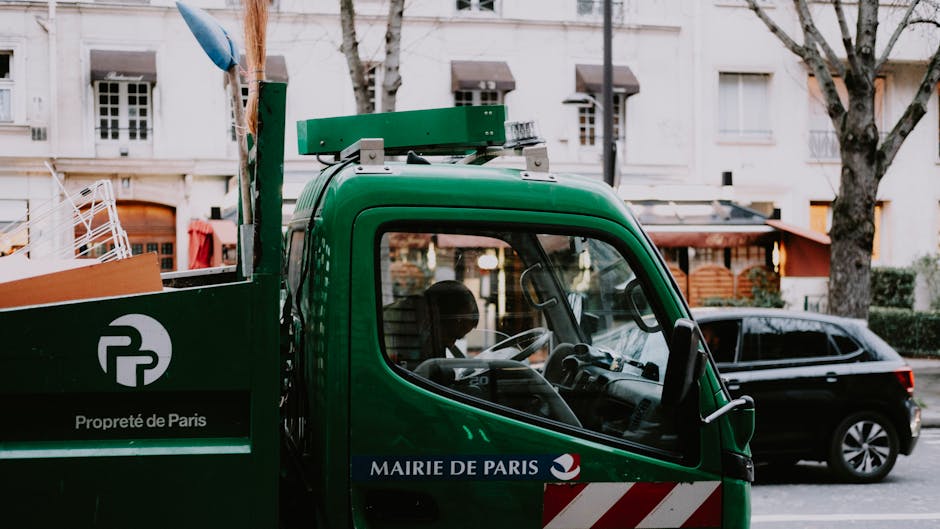 Close-up of a green rubbish collection truck parked on a city street, with the driver's side window showing a driver inside. The truck bears a white circular logo with the text 'Propreté de Paris' and a blue banner with the words 'MAIRIE DE PARIS.' In the background, there are leafless trees and a white multi-story building with rectangular windows, some open, and small balconies. A dark-colored station wagon is parked parallel to the truck, and part of a pedestrian sidewalk with a pole and a tree is visible to the left. The scene appears to be during daytime, with natural light illuminating the urban environment, reflecting the typical setting of private waste collection in a city area involving waste management services like Rubbish Clearance Marylebone's rubbish removal solutions.