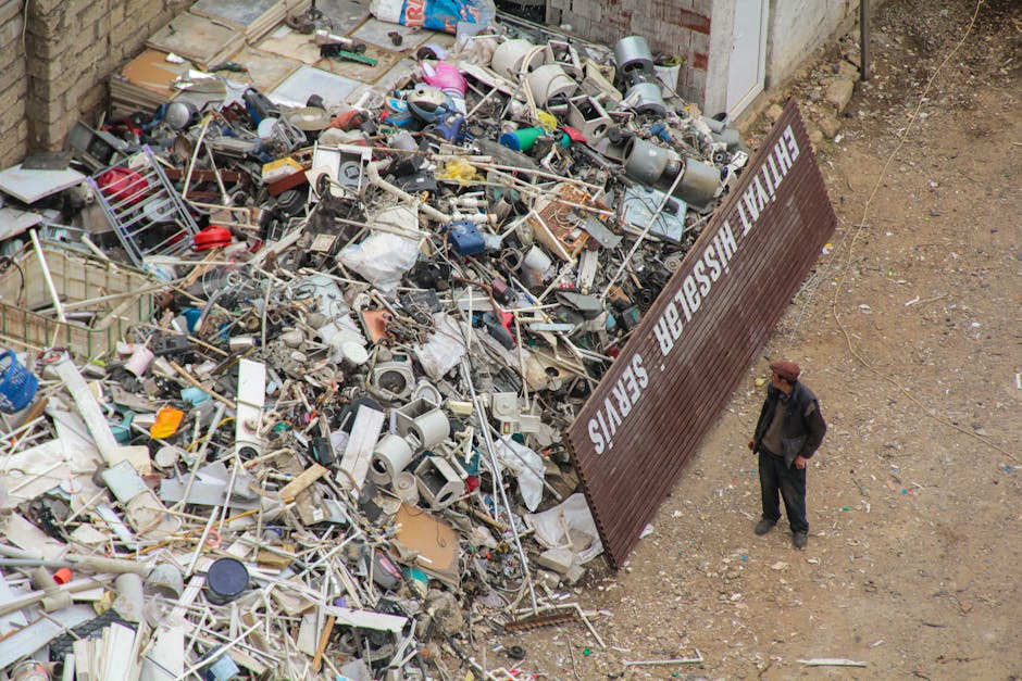 A large pile of mixed waste materials, including broken appliances such as kettles and small household items, situated outdoors on a gravel surface next to a brick wall and a signboard that reads 'RECYCLER' and 'HEALTHY WASTE SERVICES'. The debris comprises plastic, metal, and electrical components, with some items partially crushed or disassembled. On the right side of the image, a man wearing a dark jacket and a brown hat stands observing the rubbish heap, providing a sense of scale to the accumulation. The scene appears to be part of an area designated for waste collection or disposal, with the clutter suggesting the need for professional rubbish removal or alternative waste handling services commonly used for large or bulk items. The environment is well-lit, indicating natural daylight, and the surroundings reveal bare ground and a partly visible wall, typical of an outdoor waste storage or transfer site. This image effectively illustrates the kind of waste accumulation that companies like Rubbish Clearance Marylebone might handle through private collection or on-site clearance of bulky items and general rubbish.