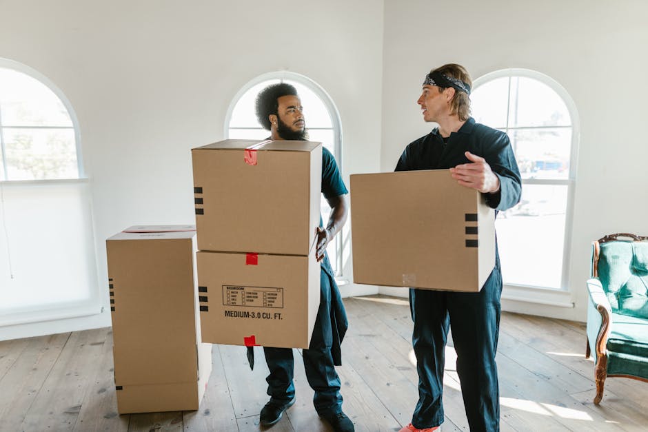 Two men are engaged in an indoor scene, each holding cardboard boxes, with one man on the left having curly black hair and a beard, and the other on the right with straight light brown hair and wearing a black headband. The man on the left is holding two stacked cardboard boxes, one labeled 'medium 3.0 cu. ft.', while the man on the right holds a single box. The room has white walls and large, arched windows allowing natural light to brighten the space. The flooring is wooden with a natural finish, and in the background, there is a green upholstered armchair with a carved wooden frame positioned near the window. There are additional cardboard boxes stacked on the floor to the left, suggesting a packing or moving activity associated with home decluttering or furniture removal services. This scene aligns with a professional rubbish removal or clearance context, highlighting aspects of independent disposal or on-site clearance as part of private waste management solutions provided by Rubbish Clearance Marylebone.