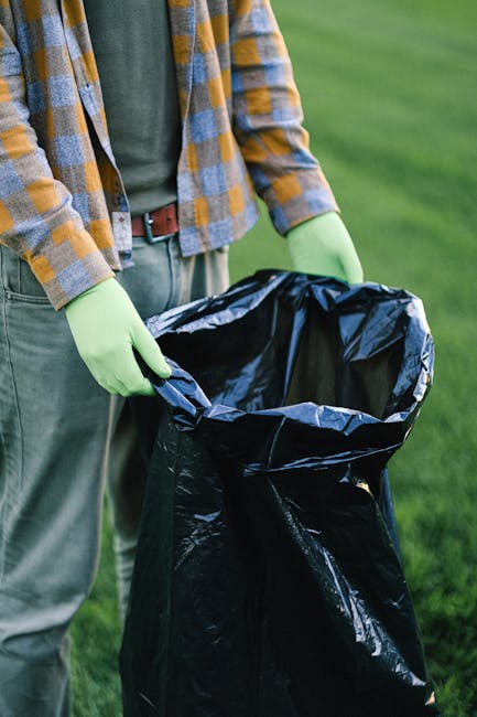 A person wearing a plaid flannel shirt in shades of yellow, grey, and blue, along with a green long-sleeved top and grey trousers, is holding the open top of a large black rubbish bag lined with a silver inner layer, with their hands covered in light green disposable gloves. The scene is set outdoors on a grassy area, which appears to be a park or garden, with vibrant green grass visible in the background. The individual is preparing to dispose of waste, possibly as part of a rubbish removal or on-site clearance activity. The black rubbish bag is empty but ready to be filled with debris or waste materials, and the overall setting suggests an informal waste management process, such as cleaning up a garden, park, or outdoor space. The image subtly reflects the process of independent waste disposal, aligned with private rubbish removal services provided by companies like Rubbish Clearance Marylebone.