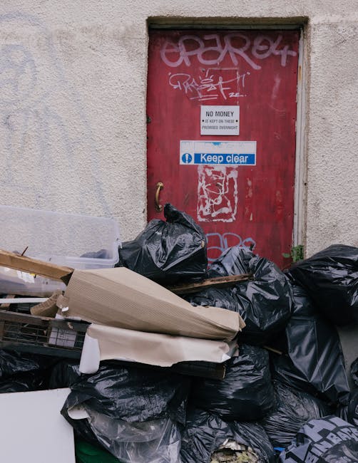 A pile of black rubbish bags filled with waste material is stacked against a beige, textured exterior wall, partially covering a red metal door that has graffiti tags and stickers. The bags are tied at the top, and some appear slightly bulging, indicating they are full. Among the waste, there are visible flattened cardboard boxes and pieces of discarded packaging, suggesting a recent or ongoing rubbish clearance. The door displays a white sign with black text indicating 'No Money' and 'Keep Clear,' along with a blue sticker that reads 'Keep clear.' The environmental setting appears to be an outdoor urban area, possibly a rear alley or service entrance, where private or independent waste disposal might take place rather than official municipal collection. The photograph captures a scene reflecting informal rubbish handling, with waste awaiting removal or collection, consistent with the context of alternative waste management and private rubbish clearance services such as those offered by Rubbish Clearance Marylebone.