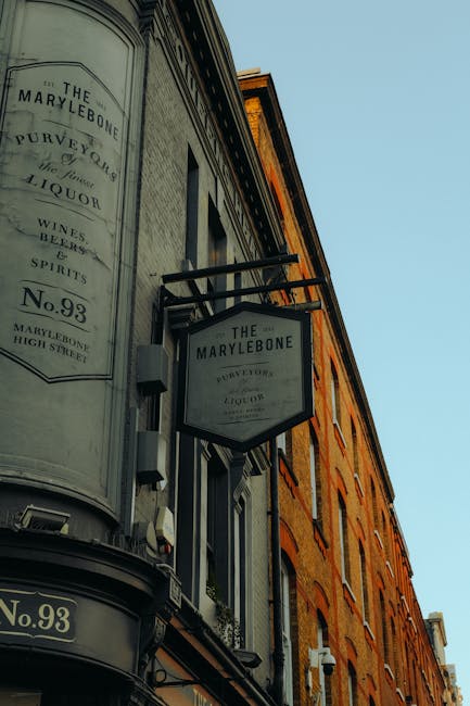 A street-level view of a building facade showing signs for 'The Marylebone,' a venue located on High Street in Marylebone. The signage is mounted on the corner of the building, with one sign hanging from a black metal bracket and another painted on a curved, light-colored surface. The signs display the venue's name, address, and details such as 'wines, beers, spirits.' The building has a classic brick exterior with a warm, reddish-brown finish, featuring several evenly spaced windows with black frames and white window surrounds. The sky in the background is clear and blue, providing natural daylight that illuminates the brickwork and signage. The scene is free of visible rubbish or waste materials, but it reflects an environment where private or independent venue management may involve on-site waste handling and rubbish clearance activities. The image suggests a typical urban setting, potentially supported by local waste services like Rubbish Clearance Marylebone, for efficient rubbish removal in commercial or residential areas.
