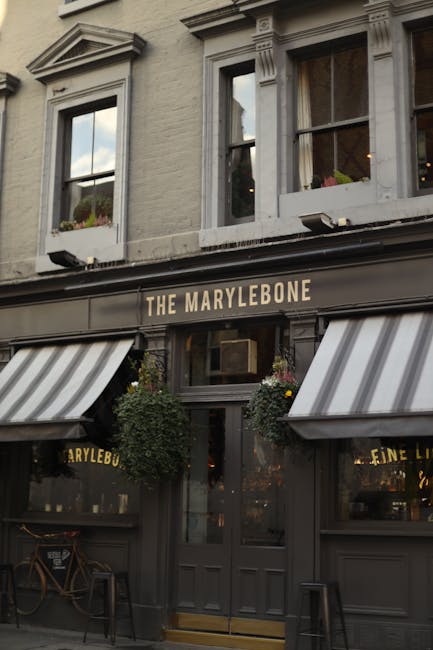 The exterior façade of a building housing 'The Marylebone,' a pub or restaurant, features a dark grey storefront with a sign displaying the establishment's name in capital letters. Above the entrance, there are two tall, narrow windows with white trim, each containing potted plants on the sills. The ground floor is adorned with two striped fabric awnings in white and grey, partially extending over the sidewalk. The entrance door is centered, made of dark wood with a glass pane, flanked by two hanging baskets filled with greenery and flowers. To the left of the door, a bicycle is parked against the wall, and to the right, there is a small stool or bench. The scene is illuminated by natural daylight, reflecting a typical urban streetscape aligned with local independent venues that might benefit from private waste handling services like rubbish clearance. The building’s exterior is well-maintained, with decorative architectural elements such as molding and trim, emphasizing its traditional, inviting appearance within a busy city environment associated with efficient rubbish removal solutions by companies like Rubbish Clearance Marylebone.