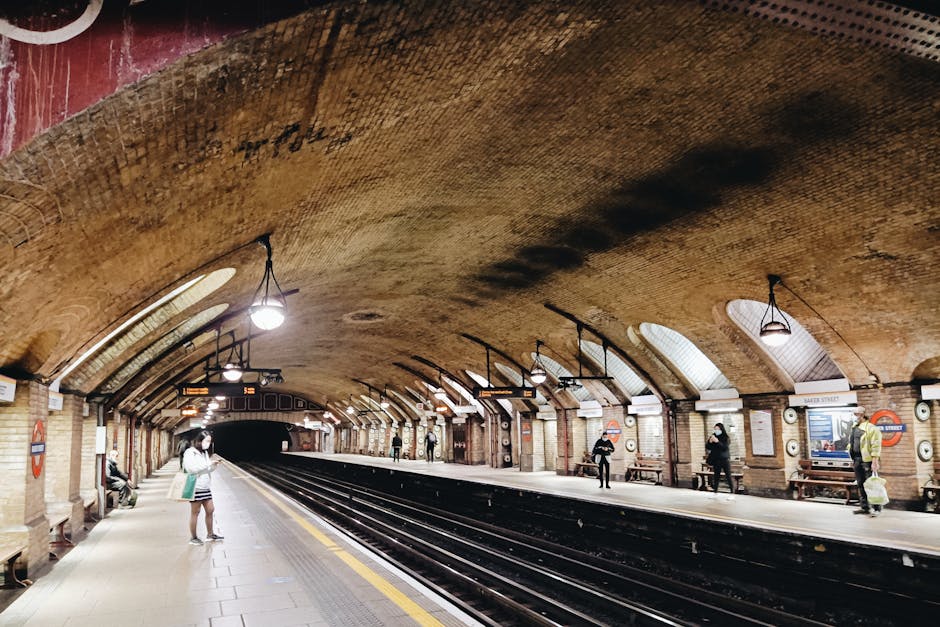 Inside a subterranean London Underground station with a curved brick arch ceiling and vintage-style lighting fixtures hanging from the ceiling. The platform is lined with wooden benches, and several passengers are waiting, some standing and others sitting. One person near the foreground is wearing a face mask and holding a green bag, while others are engaged in their own activities or looking towards the dark tunnel ahead. The station features classic signage, including roundel signs displaying the London Underground logo and station names, with one visible sign reading 'BAKER STREET.' The platform edges are marked with yellow safety lines, and the overall environment has a warm, historic atmosphere. The setting relates to private or alternative transportation options, such as independent travel arrangements, and aligns with services like rubbish clearance that may involve logistical planning in urban infrastructure environments.
