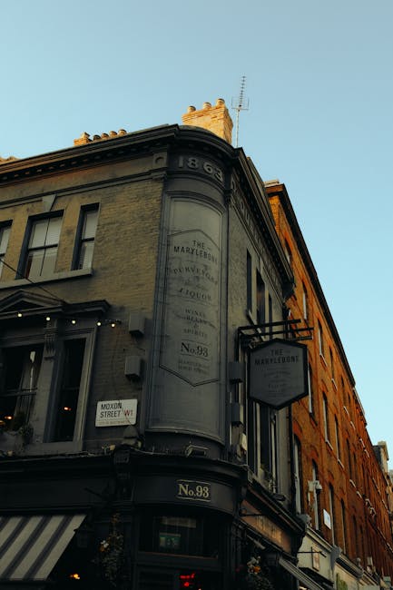 The exterior façade of a building housing 'The Marylebone,' a pub or restaurant, features a dark grey storefront with a sign displaying the establishment's name in capital letters. Above the entrance, there are two tall, narrow windows with white trim, each containing potted plants on the sills. The ground floor is adorned with two striped fabric awnings in white and grey, partially extending over the sidewalk. The entrance door is centered, made of dark wood with a glass pane, flanked by two hanging baskets filled with greenery and flowers. To the left of the door, a bicycle is parked against the wall, and to the right, there is a small stool or bench. The scene is illuminated by natural daylight, reflecting a typical urban streetscape aligned with local independent venues that might benefit from private waste handling services like rubbish clearance. The building’s exterior is well-maintained, with decorative architectural elements such as molding and trim, emphasizing its traditional, inviting appearance within a busy city environment associated with efficient rubbish removal solutions by companies like Rubbish Clearance Marylebone.