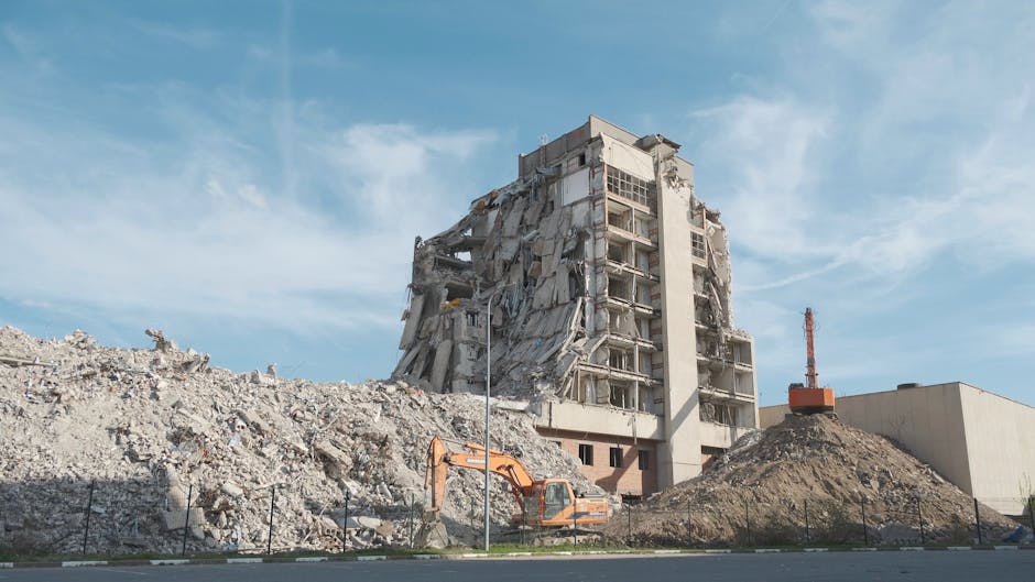 The image depicts a partially demolished multi-story concrete building with exposed floors and damaged exterior walls, situated in an open area with a clear blue sky in the background. Piles of rubble and debris surround the building's foundation, consisting of broken concrete, bricks, and dust. An orange excavator with a bucket attachment is positioned on the ground near the rubble, indicating active demolition work. A temporary fence made of wire mesh runs along the perimeter of the site. The scene appears to be an ongoing building deconstruction or renovation project, highlighting the importance of efficient waste removal and disposal services. The overall environment remains well-lit with natural daylight, emphasizing the textures of the disturbed concrete and debris, which are typical of construction site waste often handled by private rubbish collection companies such as Rubbish Clearance Marylebone. The setting suggests an urban or semi-urban context where structured demolition waste clearance may be needed to prepare the site for future development or redevelopment, aligning with professional on-site clearance practices for construction-related rubbish disposal.