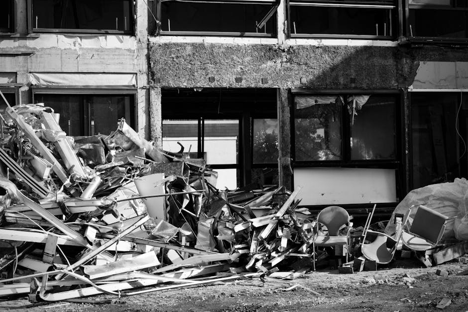 A large accumulation of metal debris and construction waste, including twisted and broken metal beams, panels, and dismantled equipment, is piled up in front of an unfinished or deconstructed building structure. The debris appears to be scrap metal with varying textures, some smooth and shiny, others rough and jagged, in monochrome shades of grey. Behind the pile, the building features exposed concrete walls with visible patches of rough, unfinished surfaces, and numerous windows with no glass, framed by dark metal. The ground in the foreground is uneven and covered with dirt, giving an impression of ongoing demolition or renovation work. There is a single plastic chair partially buried among the debris, and a large bag or bundle covered with a plastic sheet to the side, suggesting disposal or temporary storage of waste materials. The lighting is subdued, emphasizing the stark and gritty environment typical of a site undergoing private debris clearance, supported by an independent rubbish collection service such as Rubbish Clearance Marylebone, matching the context of alternative waste handling scenarios near Regent's Park, Marylebone.