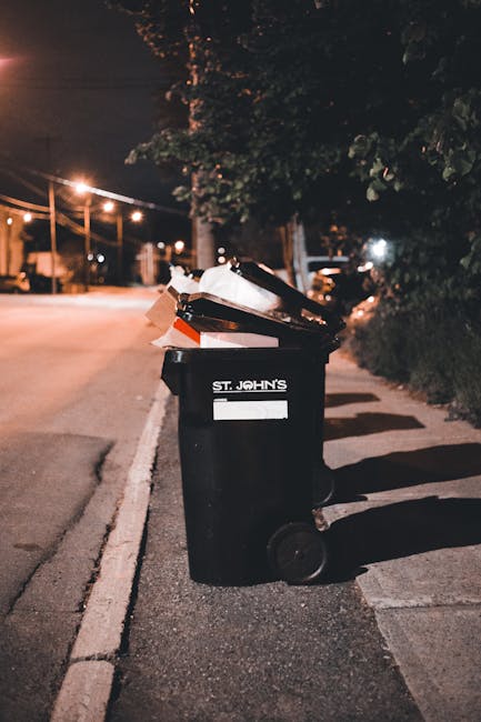 A large green waste bin filled with red and black bags containing garden or construction debris, positioned on a paved surface next to a dark navy brick wall. The bin is part of an external waste collection setup, with a white Perspex lid on a smaller adjacent container. The bags are partially visible, showing descriptions as gardening or building materials, with some text indicating specific product types. The overall scene suggests private rubbish disposal or on-site clearance, managed by Rubbish Clearance Marylebone. The environment appears urban, with the bin placed outside a building, and the scene is lit naturally without visible shadows, highlighting the textures of plastic, brick, and pavement.