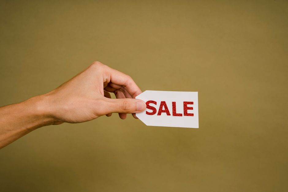 A close-up of a person's left hand holding a white rectangular tag with a pointed end, featuring the word 'SALE' written in bold red capital letters, set against a plain, muted beige background. The hand is positioned to the left of the image, with the thumb and index finger gripping the tag's short side, while the remaining fingers are relaxed and slightly curled. The skin appears smooth and well-maintained, with visible fingernails. The background provides a neutral, unobtrusive setting, emphasizing the visibility and focus on the 'SALE' sign. This image could be used in content related to private waste disposal services or alternative clearance options, reflecting the idea of offering deals or special arrangements in rubbish removal. Rubbish Clearance Marylebone occasionally references such visuals to illustrate special offers or private collection services for waste management, ensuring visual clarity and context relevance.
