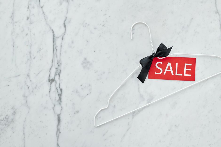 A white wire clothing hanger with a black ribbon tied into a bow around the top hook, attached to which is a red rectangular sale tag with the word 'SALE' printed in white capital letters. The hanger is placed on a light grey, textured marble surface with visible veins and subtle natural patterning. There are no other objects or background elements visible, emphasizing the hanger and sale tag as the focal points. The lighting is even and soft, highlighting the smooth finish of the marble and the matte texture of the paper tag, creating a clean and professional appearance. This image depicts an item typically used for hanging clothes, but here it is used as a prop to display the sale sign, subtly connecting to the theme of retail or clearance events, which is relevant in the context of rubbish or clearance services offered by Rubbish Clearance Marylebone.