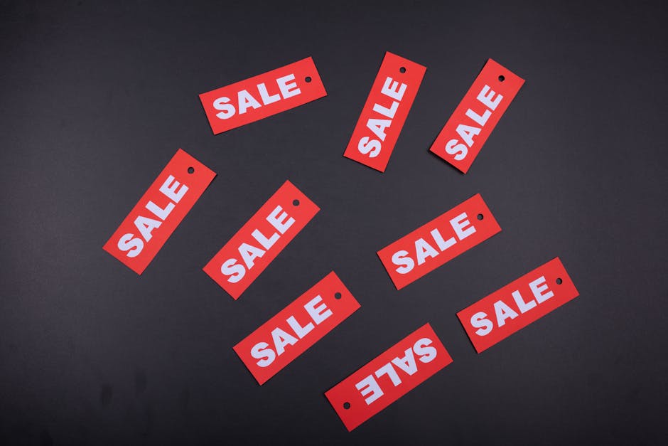 A close-up of a person's left hand holding a white rectangular tag with a pointed end, featuring the word 'SALE' written in bold red capital letters, set against a plain, muted beige background. The hand is positioned to the left of the image, with the thumb and index finger gripping the tag's short side, while the remaining fingers are relaxed and slightly curled. The skin appears smooth and well-maintained, with visible fingernails. The background provides a neutral, unobtrusive setting, emphasizing the visibility and focus on the 'SALE' sign. This image could be used in content related to private waste disposal services or alternative clearance options, reflecting the idea of offering deals or special arrangements in rubbish removal. Rubbish Clearance Marylebone occasionally references such visuals to illustrate special offers or private collection services for waste management, ensuring visual clarity and context relevance.