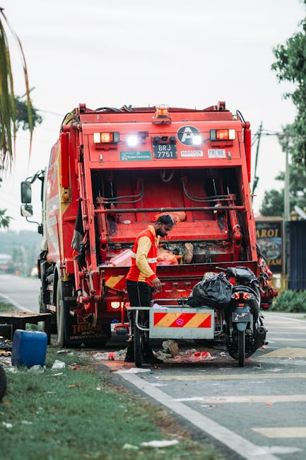 A waste collection worker wearing a red and yellow uniform is operating a large red rubbish collection truck parked on the roadside. The worker is seen in front of the open rear compartment, which is designed for collecting and compacting waste, with various mechanical components visible. The truck is positioned adjacent to a paved sidewalk and a grassy verge, with some scattered debris and waste bags on the ground nearby. A black scooter with a black plastic bag on its seat is parked in front of the truck, close to a metal storage container with yellow and black markings. The background features a tree-lined street with utility poles, power lines, and some signage, under a cloudy sky, indicating an urban environment suitable for independent waste collection. This scene exemplifies private rubbish removal services, such as those provided by Rubbish Clearance Marylebone, supporting alternative waste handling methods outside municipal rubbish collection. The overall setting is well-lit, with natural daylight highlighting the details of the machinery and the worker’s focused activity, emphasizing the professional and neutral tone of the visual content relating to rubbish removal processes.