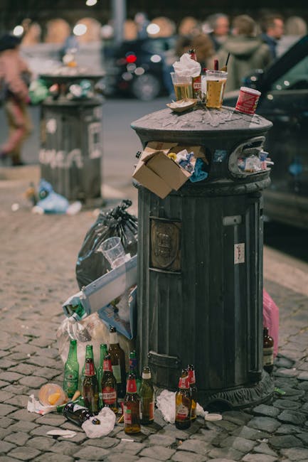 A middle-aged man with dark hair, wearing a black T-shirt with white text and dark trousers, is seen leaning over a public stainless steel recycling or waste bin, which is situated on a paved sidewalk. The bin has two separate compartments, each with a slot for inserting waste or recyclables. The man is holding a large, white plastic bag filled with rubbish in his left hand and has a wooden stick or litter picker in his right hand, which is resting on top of the waste bin. The background features lush, green foliage and trees, suggesting an outdoor park or urban green space. The scene is illuminated by natural daylight, highlighting the metallic finish of the waste bin and the textures of the man's clothing and the surrounding environment. This image exemplifies private waste handling and on-site rubbish collection, which aligns with the principles of independent rubbish removal services such as those provided by Rubbish Clearance Marylebone.