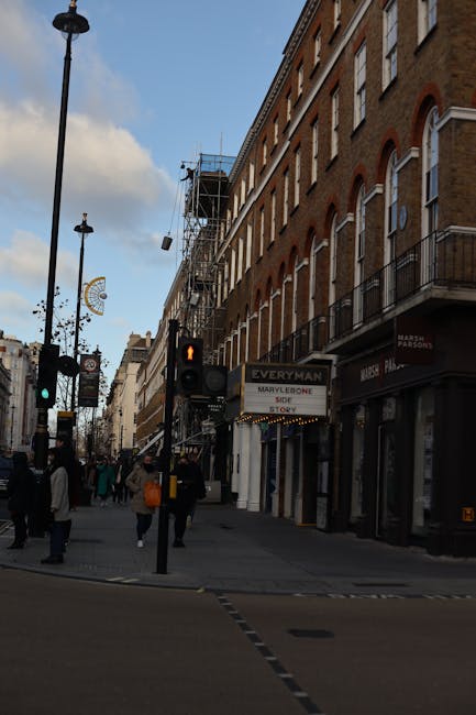 A busy street scene in an urban area shows a row of multi-storey brick buildings with large windows and small balconies. One building on the right side is under renovation, covered in scaffolding and protective netting. Pedestrians are walking along the pavement, some carrying bags or backpacks, with a few waiting to cross at a traffic light displaying an orange pedestrian signal. The street has various streetlights, and a prominent sign advertises a MarvcLove store with a smaller sign for 'Everyman' above the storefronts. In the foreground, there is a pedestrian crossing with tactile paving. The environment is illuminated by daylight, and the sky above is partly cloudy with patches of blue visible. The scene reflects a typical urban shopping district where local businesses are supported by foot traffic, and external renovation works are visible on one of the buildings, consistent with independent property maintenance by those managing or removing rubbish from site-based or private disposal projects, as handled by services like Rubbish Clearance Marylebone.