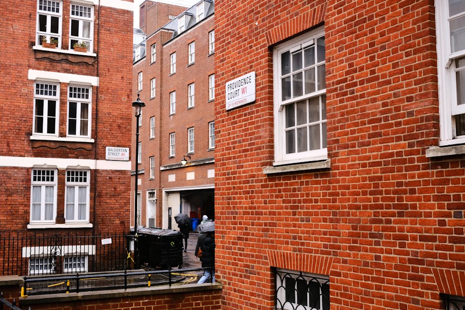 A busy street scene in an urban area shows a row of multi-storey brick buildings with large windows and small balconies. One building on the right side is under renovation, covered in scaffolding and protective netting. Pedestrians are walking along the pavement, some carrying bags or backpacks, with a few waiting to cross at a traffic light displaying an orange pedestrian signal. The street has various streetlights, and a prominent sign advertises a MarvcLove store with a smaller sign for 'Everyman' above the storefronts. In the foreground, there is a pedestrian crossing with tactile paving. The environment is illuminated by daylight, and the sky above is partly cloudy with patches of blue visible. The scene reflects a typical urban shopping district where local businesses are supported by foot traffic, and external renovation works are visible on one of the buildings, consistent with independent property maintenance by those managing or removing rubbish from site-based or private disposal projects, as handled by services like Rubbish Clearance Marylebone.