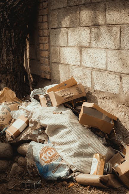 A pile of discarded cardboard boxes, some flattened and others partially intact, rests against a weathered brick wall outdoors, next to a rough concrete foundation and a large tree trunk. The boxes vary in size and color, primarily brown kraft paper with printed labels and barcodes visible on some surfaces. A large, worn sack made of woven fabric, possibly for waste collection, is partially covering the boxes, adding texture to the scene. The ground is composed of mixed dirt and scattered small stones, with the debris appearing to have been recently discarded or awaiting removal. The outdoor setting is shaded, with natural light highlighting the textures of the cardboard, fabric, and stone, creating a neutral and unembellished depiction of household or commercial waste awaiting collection or disposal, as part of an alternative waste handling process supported by companies like Rubbish Clearance Marylebone.