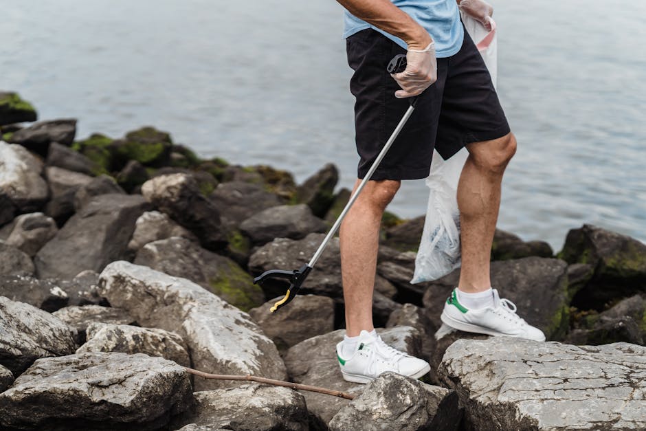 A person wearing a light blue T-shirt, black shorts, and white sneakers with green accents is standing among large, uneven rocks near a body of water. The individual is holding a long-handled grabber tool in their right hand, which is positioned slightly downward, and is wearing a white glove on that hand. The rocks are a mix of dark gray and brown tones, some with patches of green moss or algae. The water in the background appears calm, with a muted grayish-blue hue, suggesting a lakeside or seaside environment. The person's left leg is slightly bent, and they are holding a white plastic bag, which is partly visible behind their right leg. The scene portrays an outdoor setting that could be associated with shoreline cleanup or environmental conservation activities, aligning with the scope of rubbish removal and waste collection services. The lighting suggests natural daylight, casting soft shadows on the rocks and the individual's legs, emphasizing the rugged texture of the natural environment.