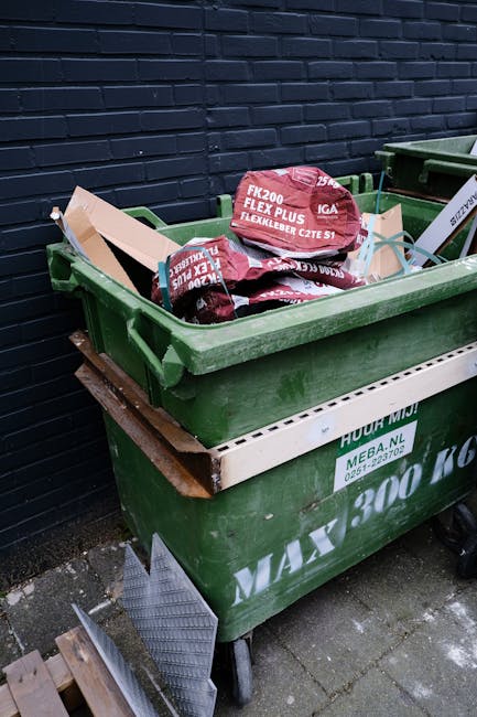 A large green wheeled skip positioned on a paved surface against a dark brick wall, filled with black and red bags of construction or garden waste, including materials labeled 'FK200 FLEXY PLUS'. A white debris containment tray is placed on the side of the skip, and adjacent to it is another green skip, suggesting a private waste collection or on-site clearance operation. The surrounding environment appears to be an outdoor urban setting, typical for rubbish removal services such as those provided by Rubbish Clearance Marylebone. The scene shows a mix of waste bags and limited visible debris, indicative of a typical waste disposal or rubbish clearance scenario before collection. Natural daylight illuminates the area, highlighting the textures and colors of the materials and the brick wall background, emphasizing its practical, functional purpose in managing rubbish in a professional waste management context.