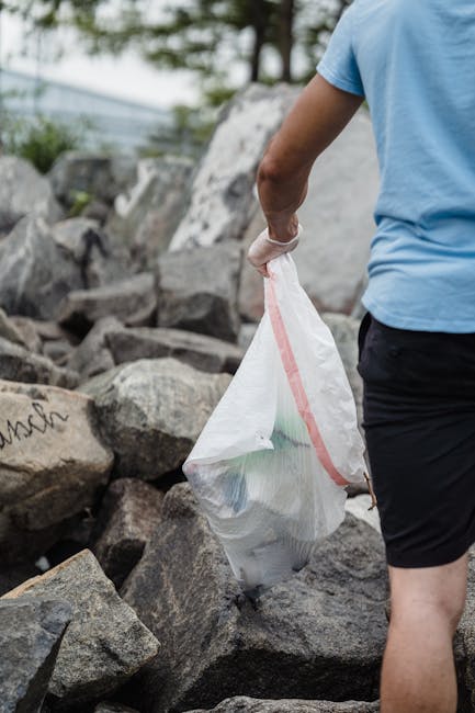 A person wearing a light blue T-shirt, black shorts, and white sneakers with green accents is standing among large, uneven rocks near a body of water. The individual is holding a long-handled grabber tool in their right hand, which is positioned slightly downward, and is wearing a white glove on that hand. The rocks are a mix of dark gray and brown tones, some with patches of green moss or algae. The water in the background appears calm, with a muted grayish-blue hue, suggesting a lakeside or seaside environment. The person's left leg is slightly bent, and they are holding a white plastic bag, which is partly visible behind their right leg. The scene portrays an outdoor setting that could be associated with shoreline cleanup or environmental conservation activities, aligning with the scope of rubbish removal and waste collection services. The lighting suggests natural daylight, casting soft shadows on the rocks and the individual's legs, emphasizing the rugged texture of the natural environment.