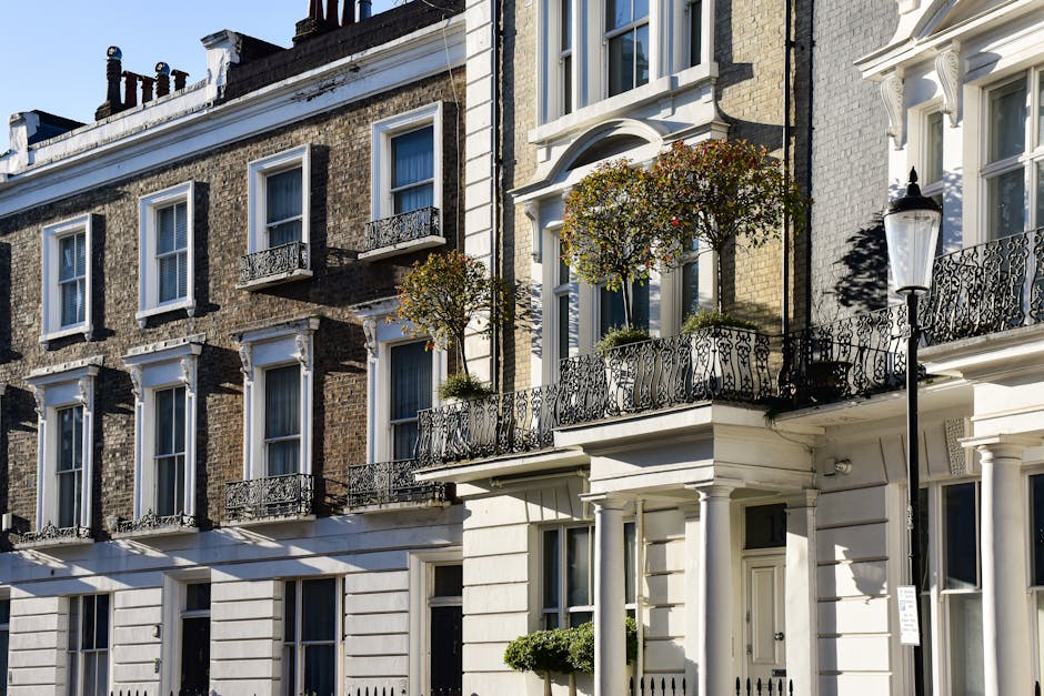 A row of Victorian-style terraced townhouses with a combination of brick and white-painted facades, featuring decorative white cornices and window trims. Each property has multiple sash windows with white frames, and some have small wrought iron balconies with intricate black railings. The buildings are four stories tall, and the balconies on the upper floors are adorned with potted plants and small trees, adding greenery to the scene. To the right, a traditional black cast-iron lamppost stands on the sidewalk, casting a shadow onto the white facade below. The environment appears to be a residential street with paved pavements and limited street furniture. The lighting suggests a clear, sunny day, with sharp shadows indicating direct sunlight. This scene shows well-maintained, historic homes that may require occasional on-site or private rubbish disposal, aligning with independent waste clearance services that handle residential rubbish collections in Marylebone. Rubbish Clearance Marylebone specializes in such services, supporting local residents with waste management outside of council collection routines.